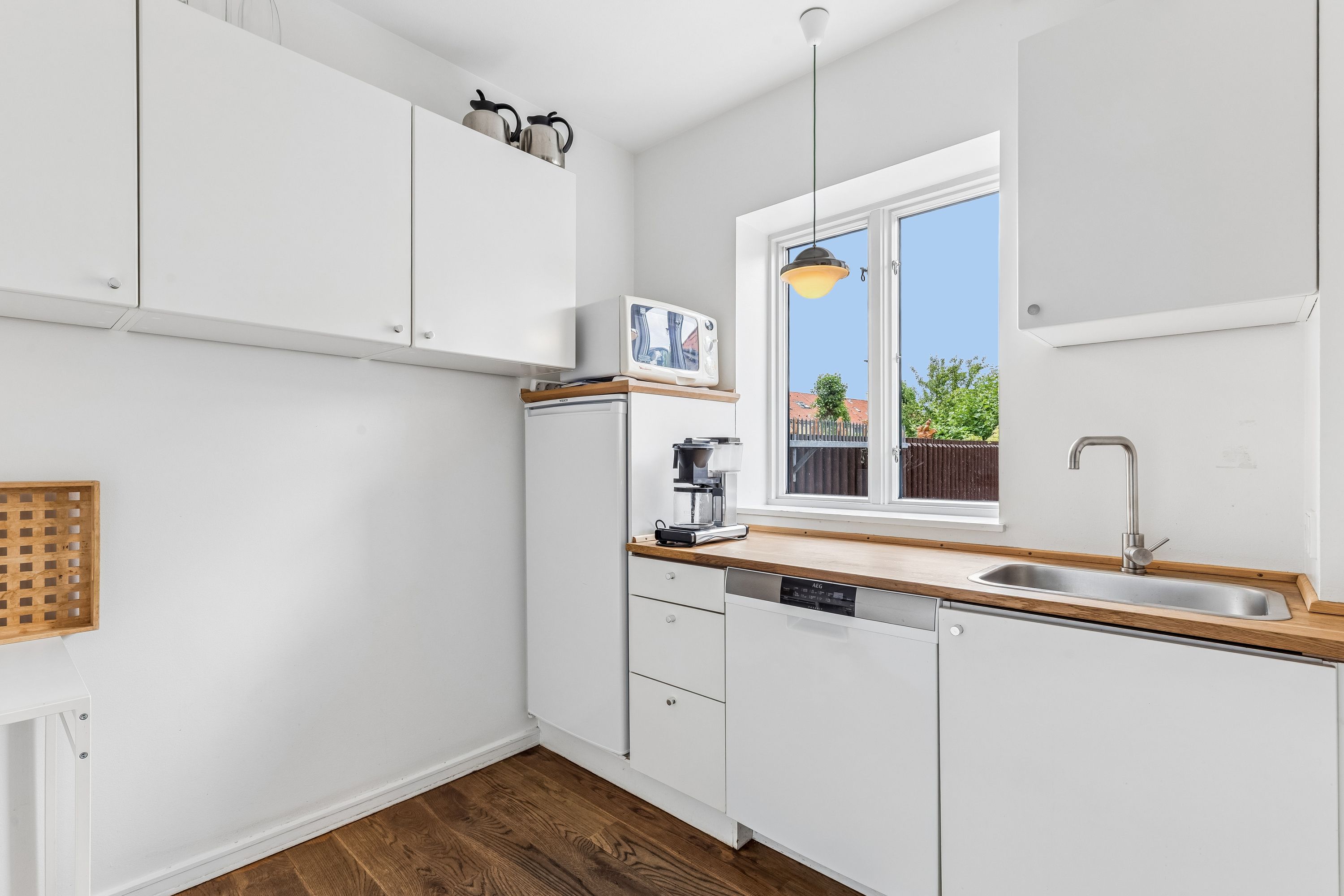 En hvid stue med trægulv, hvide vægge og lyse loft. Et hængelampe og en åben vinduesfløj. Køkken med moderne hvide indretning og trækøkkenbord. Et mikroovn og en kaffemaskine på en hylde.