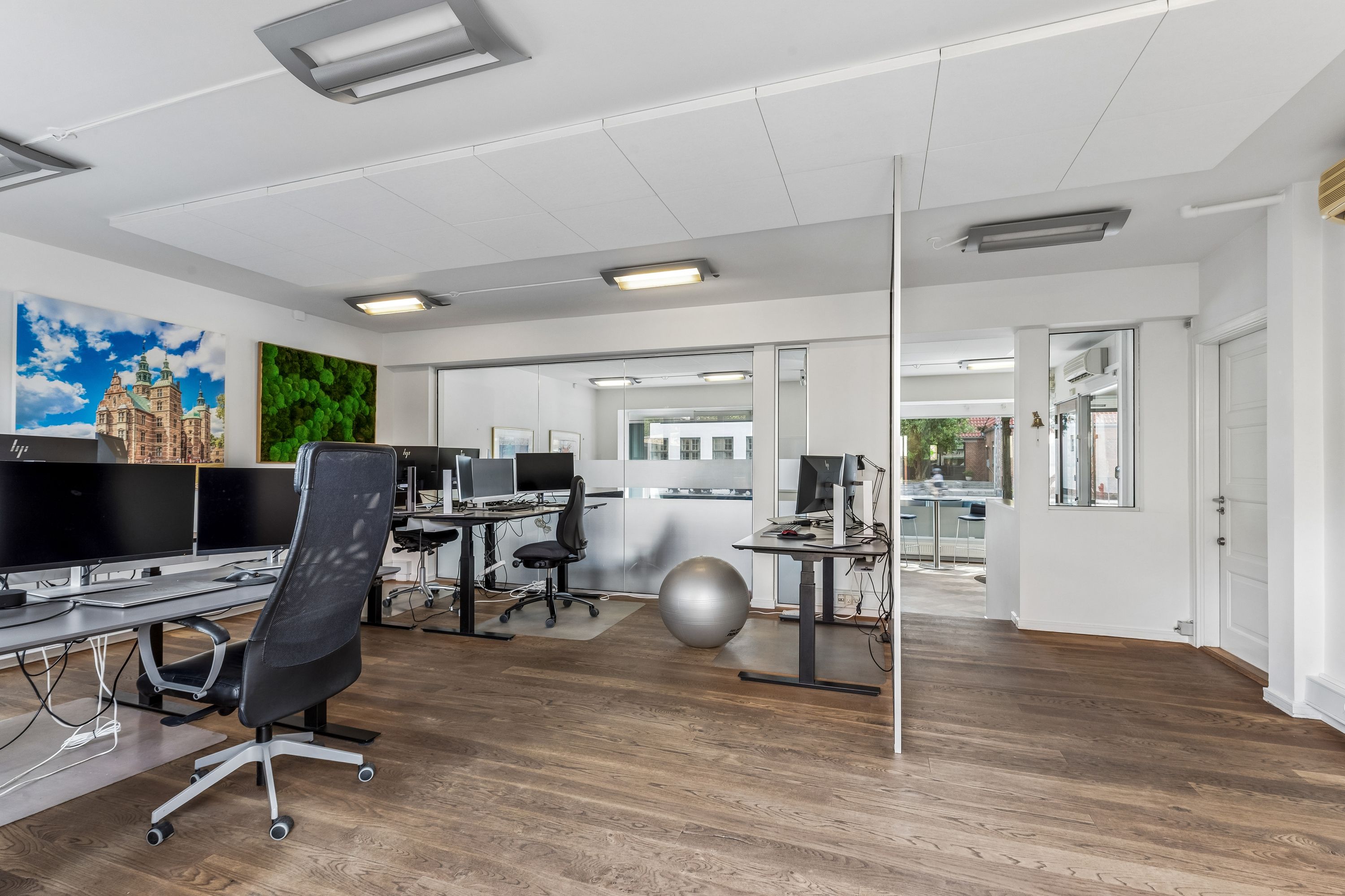 Kontor med moderne skriveborde, skærme og møbler samt et lysere loft med indbygget belysning og en hvid dør.