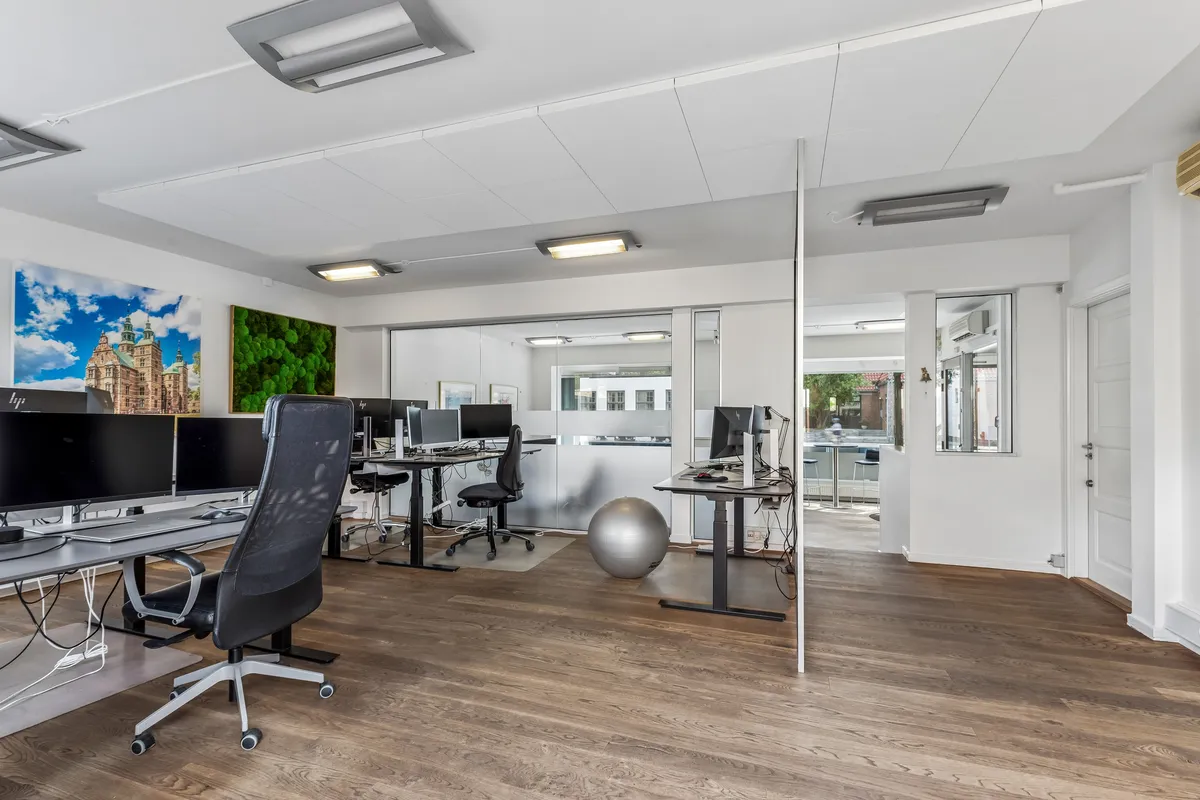 Kontor med moderne skriveborde, skærme og møbler samt et lysere loft med indbygget belysning og en hvid dør.