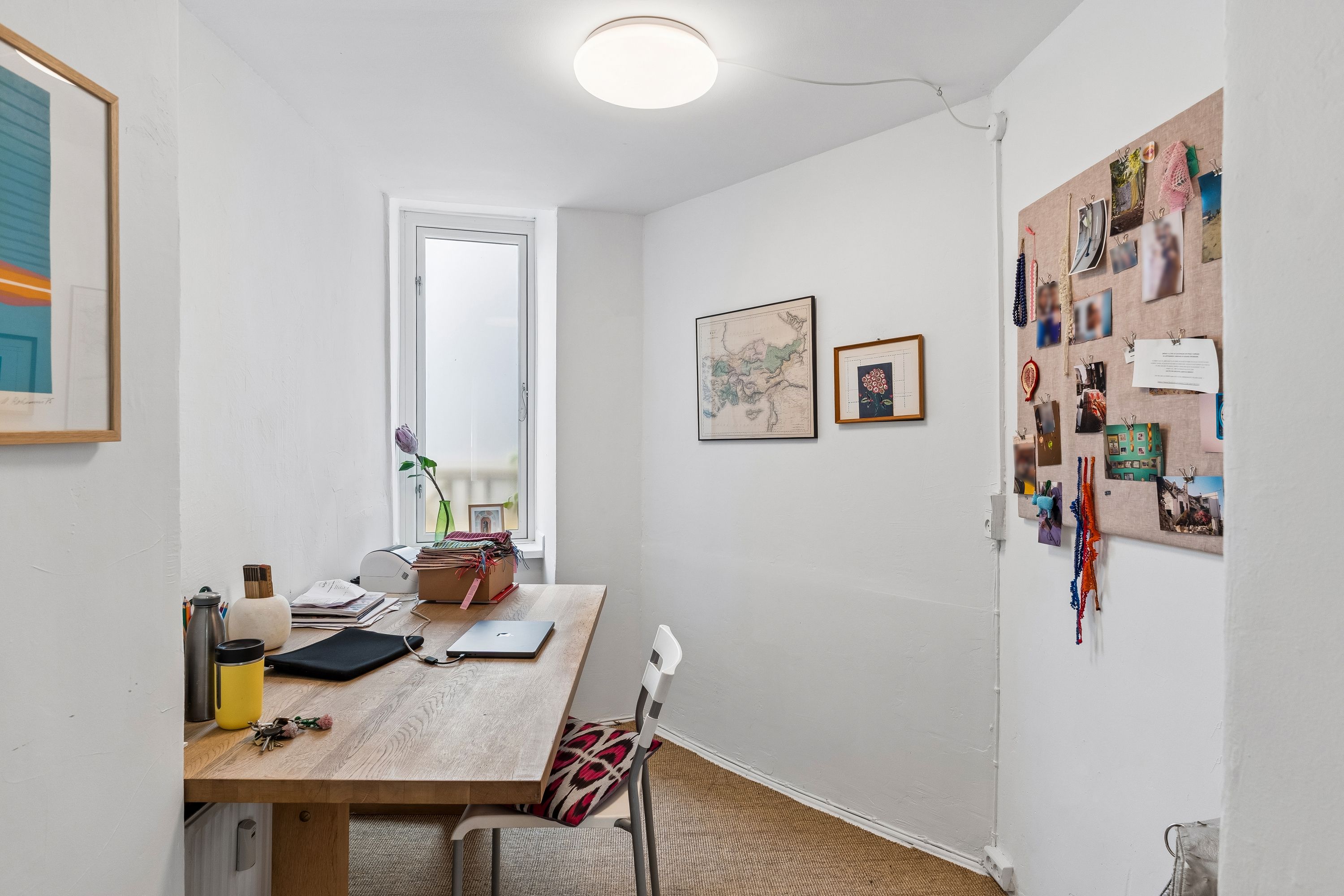 Kontor med bord, stol og en laptop, der er klar til brug. På bordet er der en flaske, nøgler og flere papirer. Et vindue giver lys ind i værelset