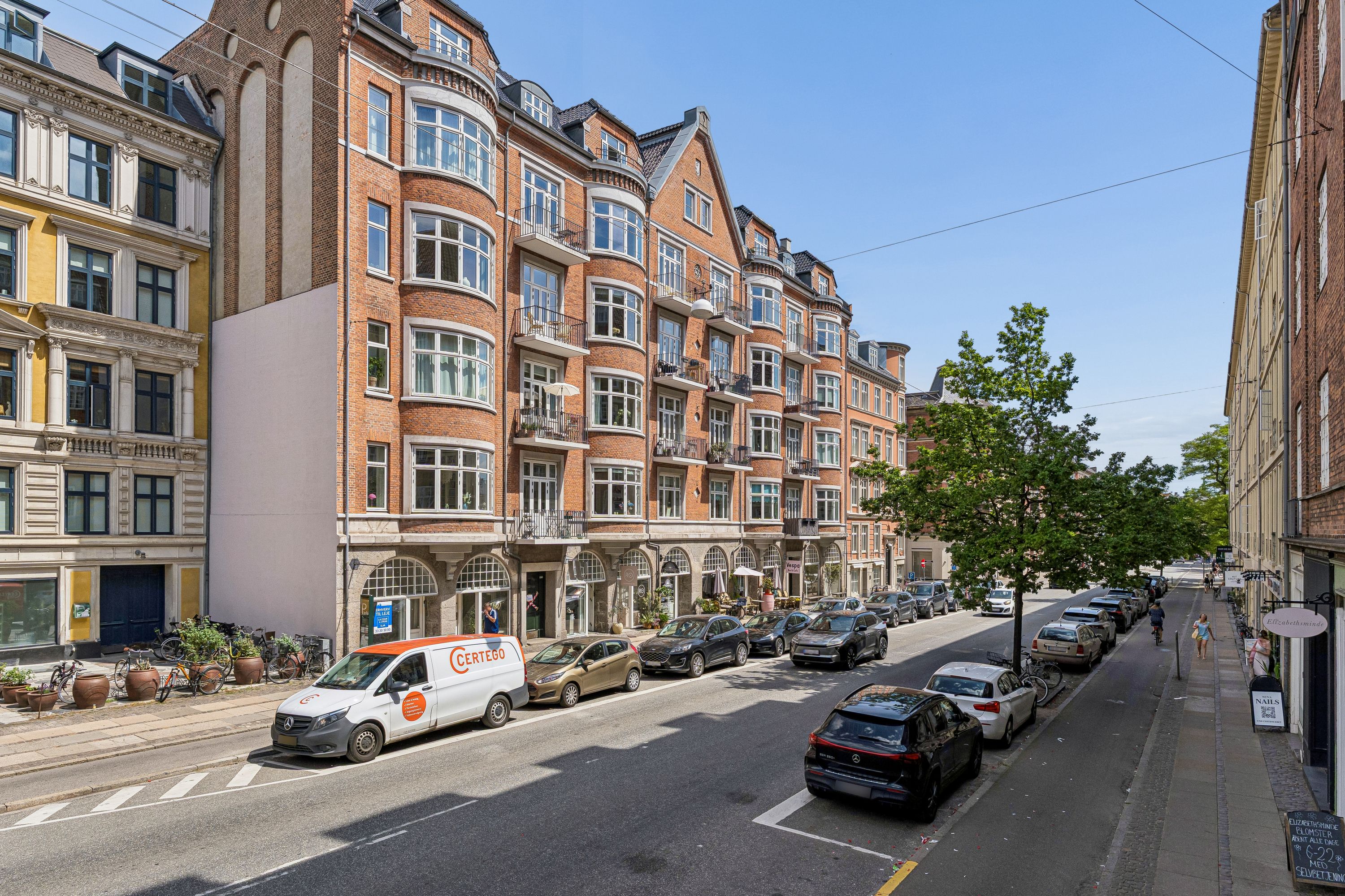 Store, røde lejlighedsbygninger på Vesterbrogade 180. Butikker med grønne planter og mennesker på fortovet. Biler parkeret langs gaden. En gul Mercedes Vito er i gang.