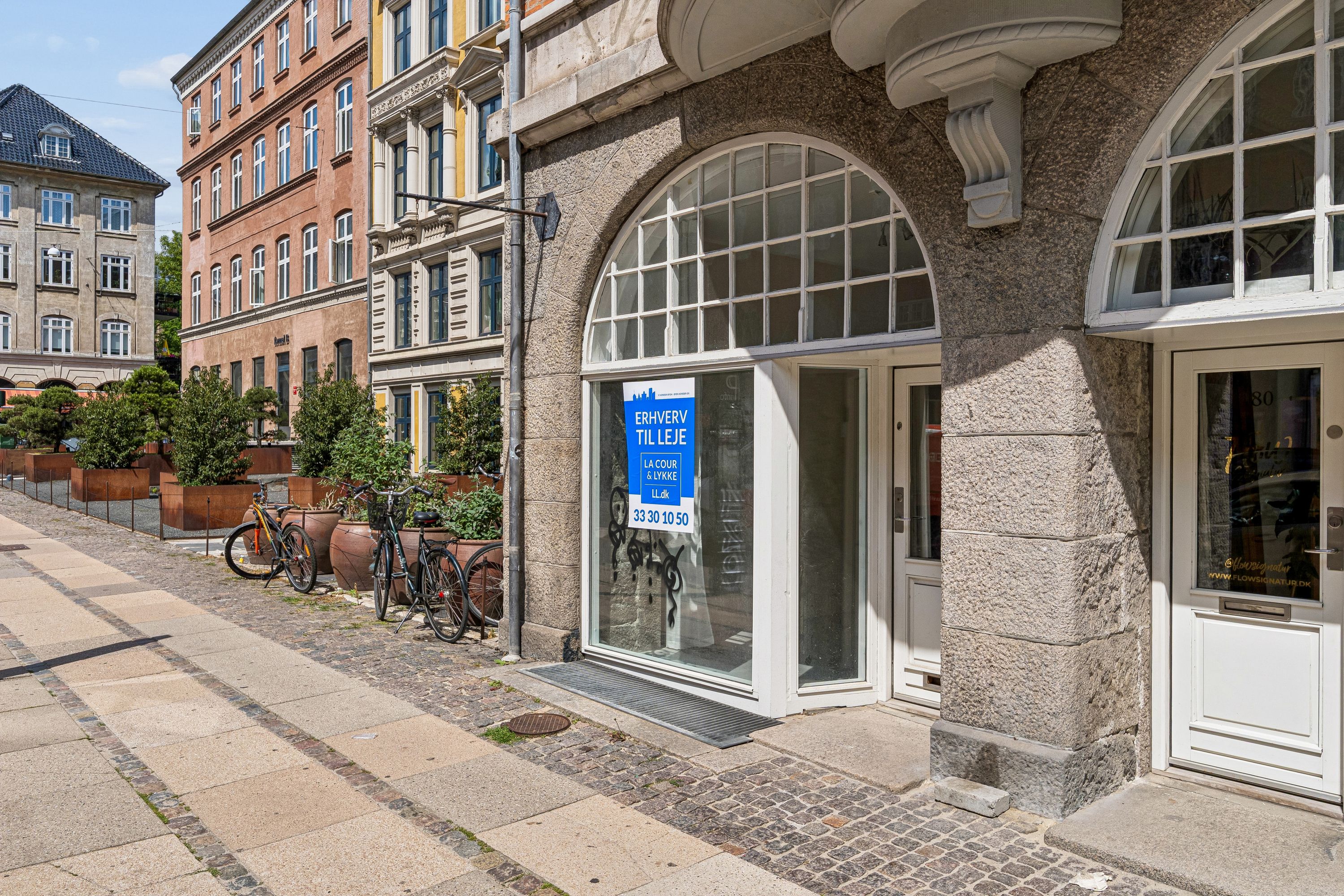 Butik med skilt, der læser Erhverv til leje. Hjørnebutik med glassdøre og vindue samt cykler og planter. Københavnsgade med betonbrolæg og bygninger i baggrunden