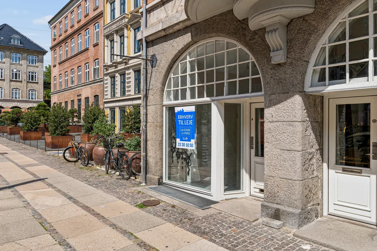 Butik med skilt, der læser Erhverv til leje. Hjørnebutik med glassdøre og vindue samt cykler og planter. Københavnsgade med betonbrolæg og bygninger i baggrunden