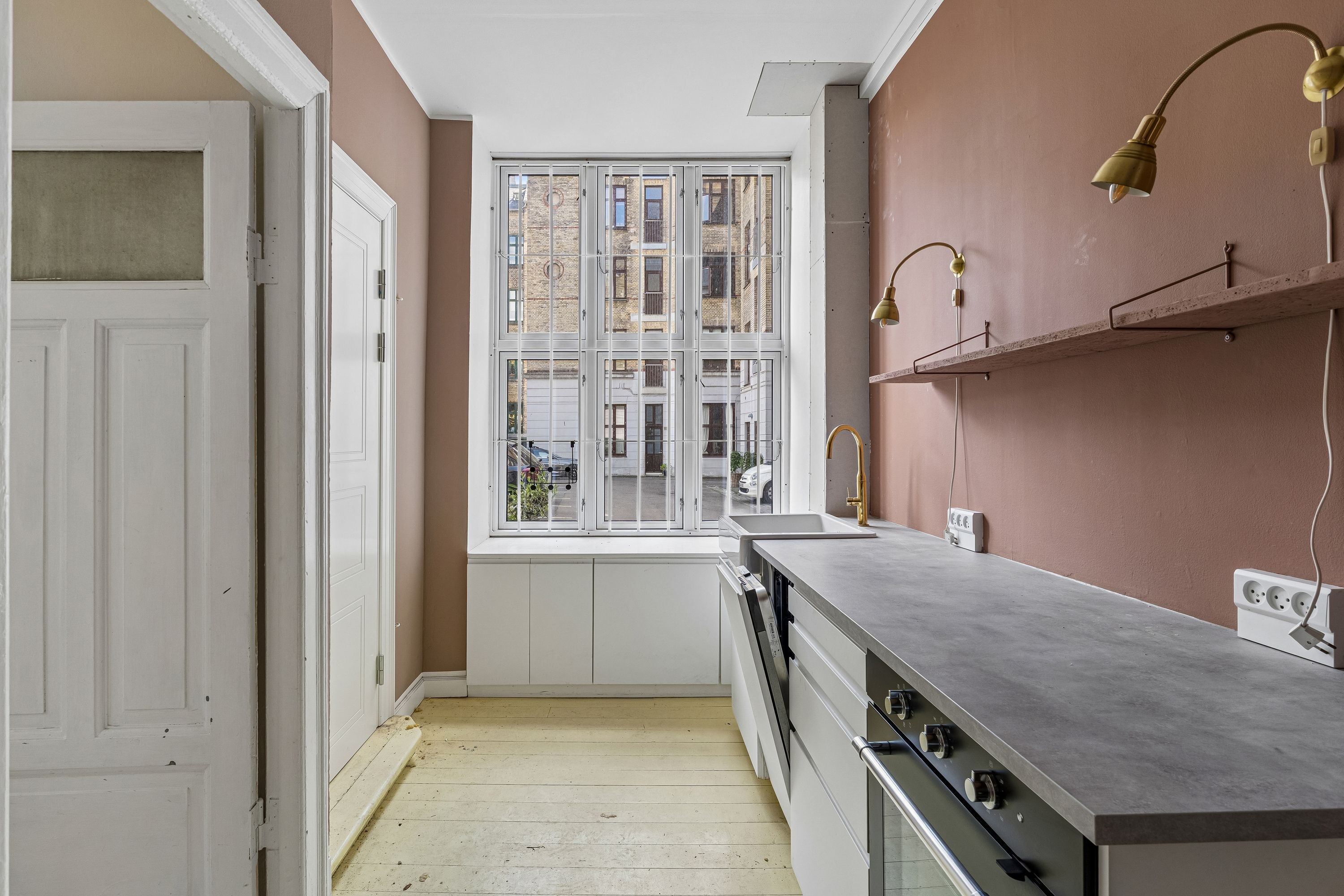 Køkken med lyse beige vægge og lysegrå loft. Hvidt køkkenbord, køkkenhøj og en ovn. To lysegule lampekroner og lysegrå væg med to hængende hylder.