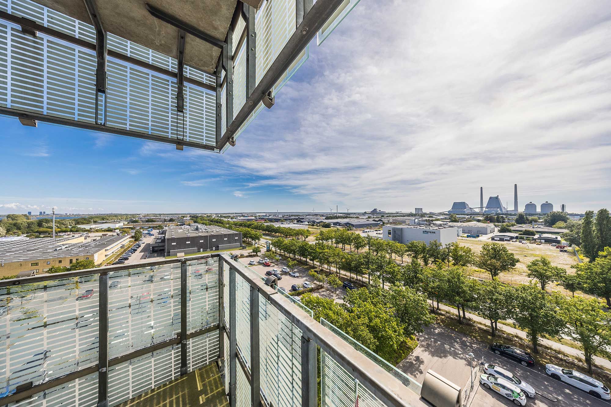 Balkon med udsigt over parkeringsplads og veje i et byområde med grønne træer og solrige himmel