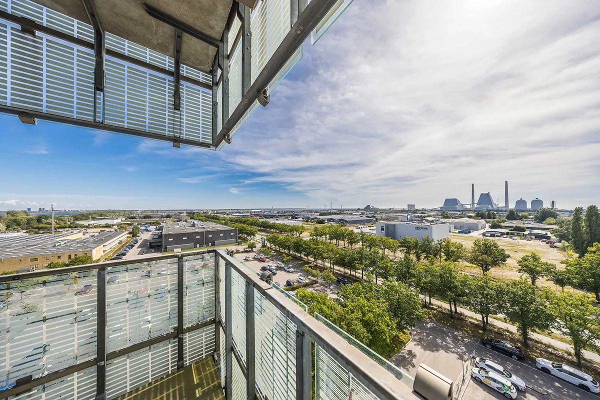 Balkon med udsigt over parkeringsplads og veje i et byområde med grønne træer og solrige himmel