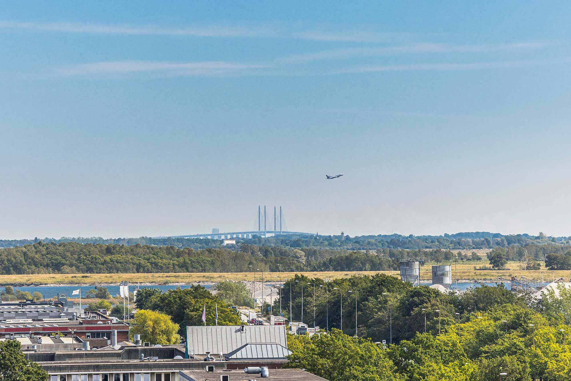 Overblik over byen med lufthavnsbroen i baggrunden og en jetfly i luften