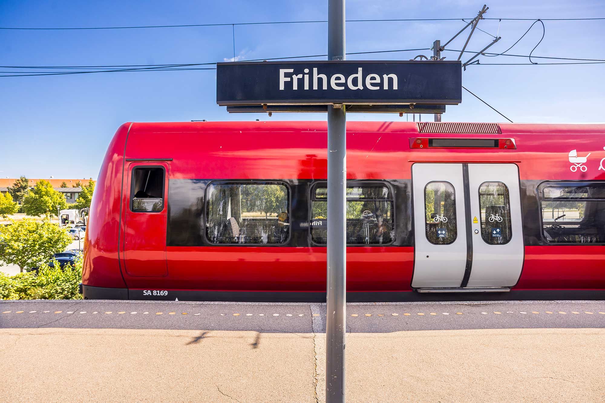 En rødt tog står på stationen Friheden med skiltet over og flere mennesker inde