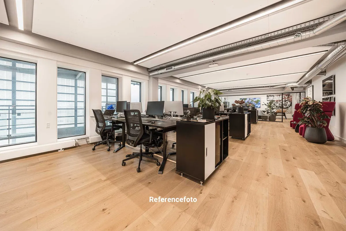 Kontor med store ruder, højt til loftet og lysegulve. Fire borde med computere og stol samt to røde lænestoler med plante.