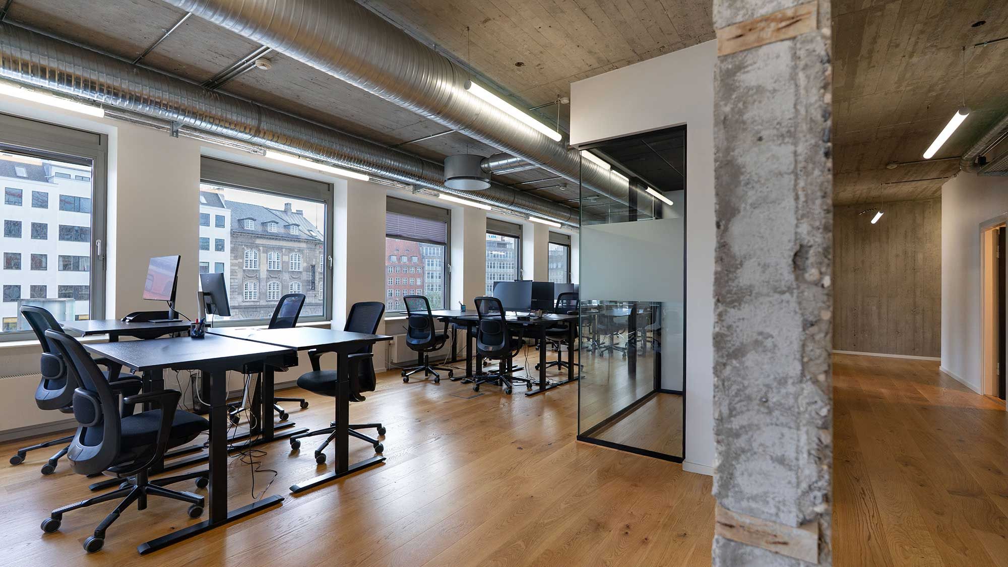 Kontor med moderne design og vinduer med udsigt til bygningerne udenfor. Rundt er der lysegul gulvbelægning og moderne møbler.