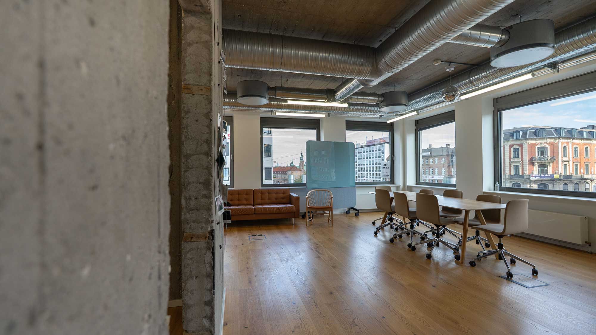 Et kontor med lyse trægulve og moderne møbler, en stol og en sofa i brun farve. Vinduerne går op mod gaderne i byen, og der er store hvide vægge med industriinspireret design.