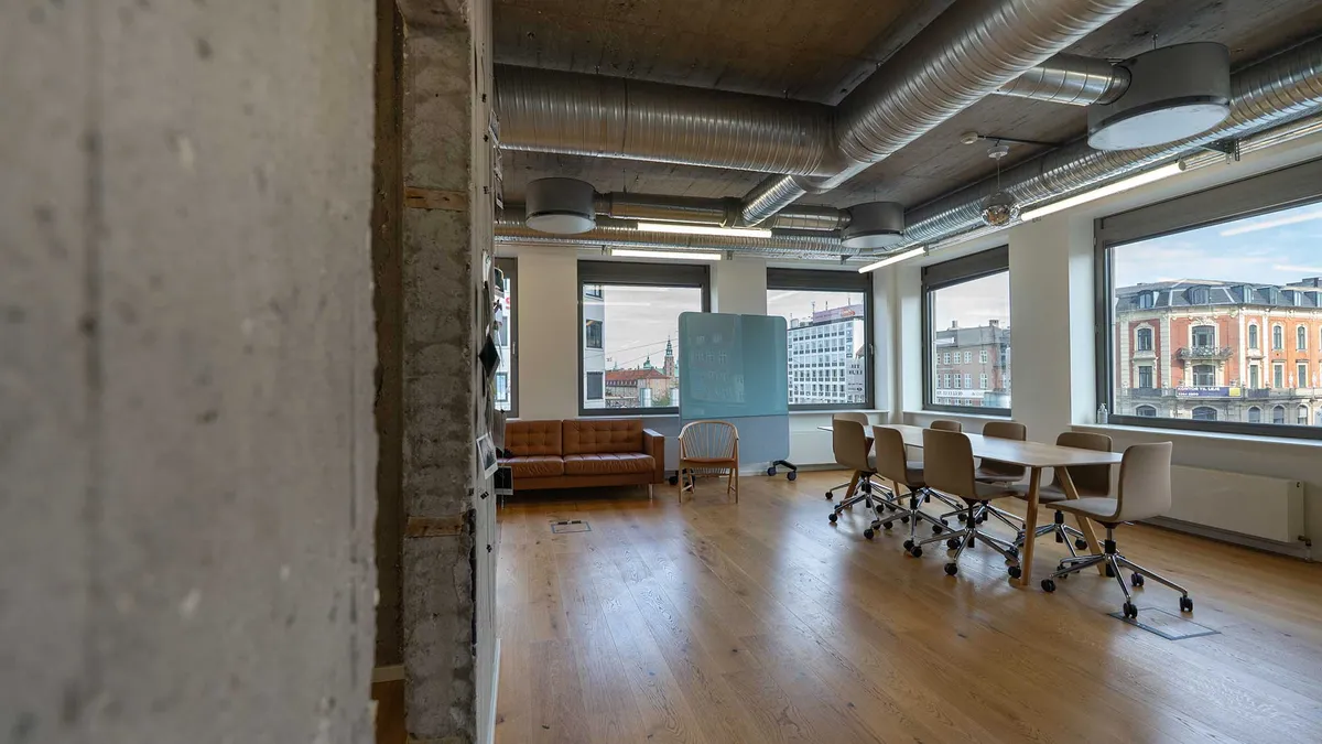 Et kontor med lyse trægulve og moderne møbler, en stol og en sofa i brun farve. Vinduerne går op mod gaderne i byen, og der er store hvide vægge med industriinspireret design.