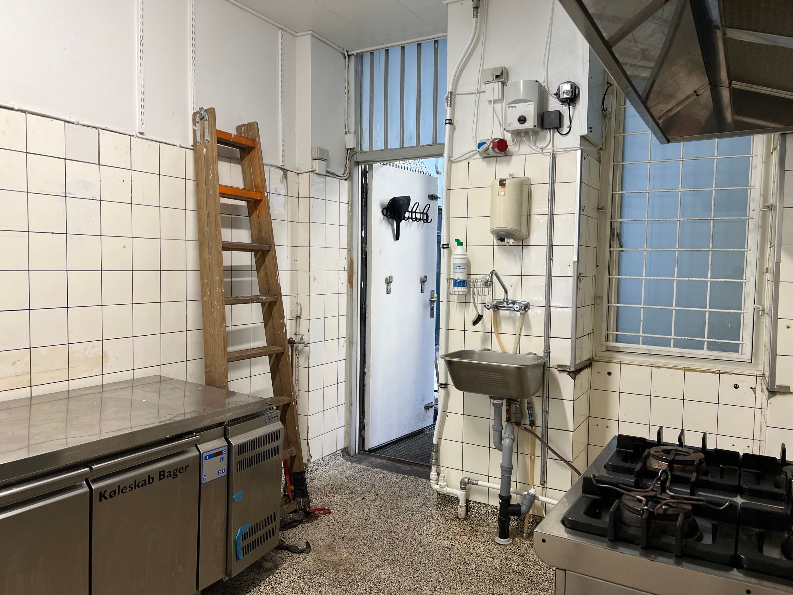 En ren og funktionel køkken i erhvervslokaler med flere ovne og skabe samt en åben dør og et vindue med glas og metal rammer