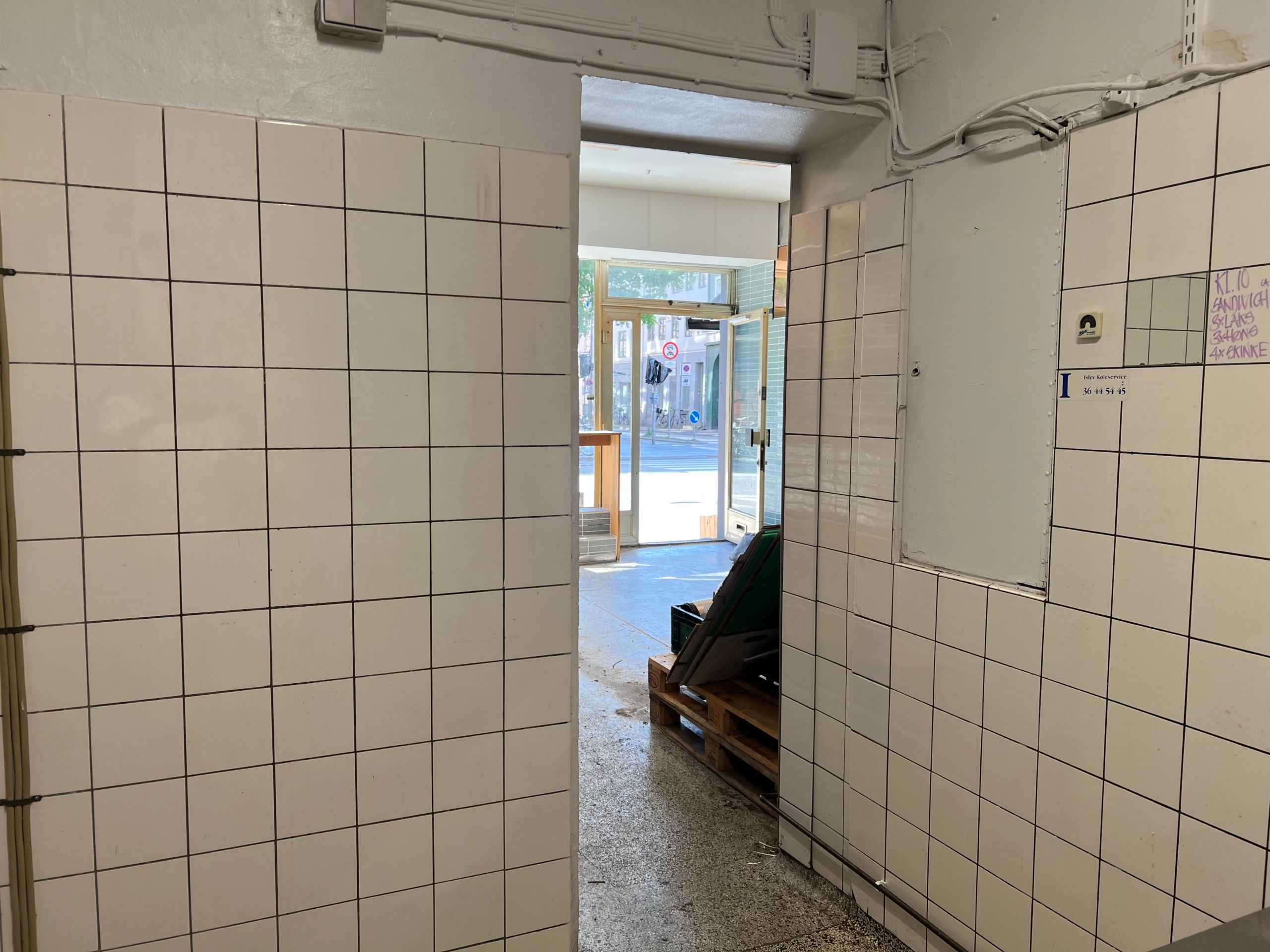 En ren køkkenafdeling med hvide fliser og tomme vægruder. En dør og et vindue er åbnet mod gaden med bygninger i baggrunden.