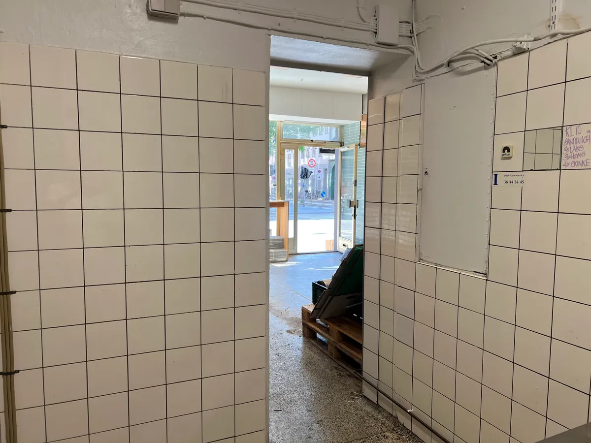 En ren køkkenafdeling med hvide fliser og tomme vægruder. En dør og et vindue er åbnet mod gaden med bygninger i baggrunden.
