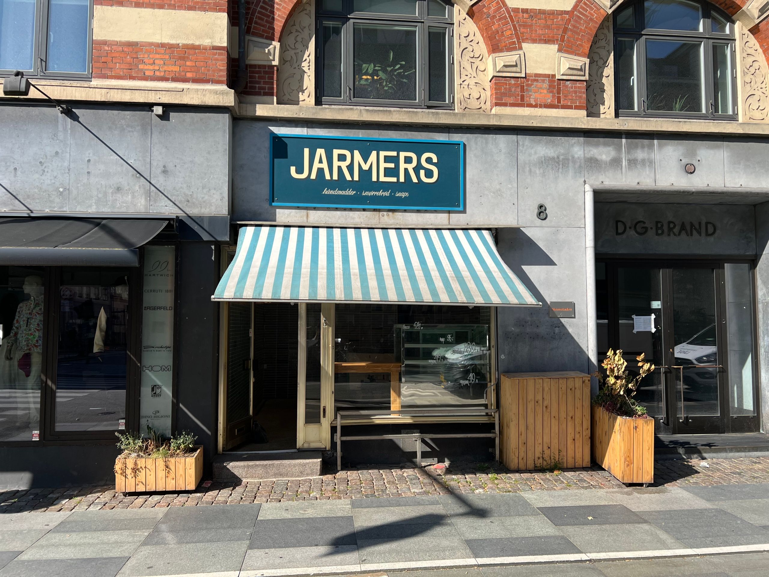 Københavnsgade med butikker og gavle, nummer 8 med butiksskilt, og et mærke for Jarmers. En gågade med pavilloner og plantepoter, og en åben dør til et lejemål