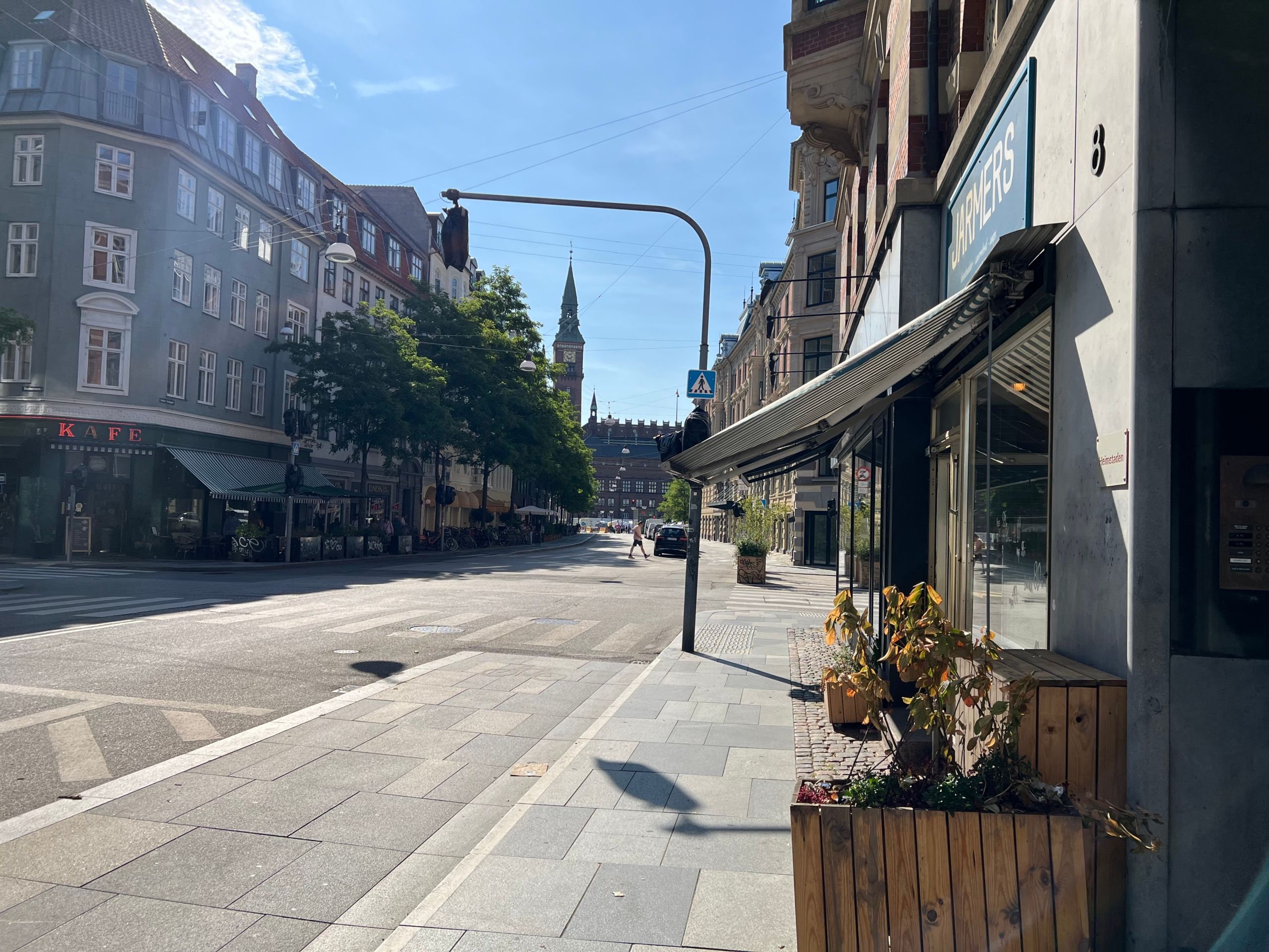Lejlighed til leje på Vester Voldgade 8. Bygningen har lyse gulve, store vinduer og en grøn have. Butikker med plantager i forgrunden. En klippe med spiret i baggrunden