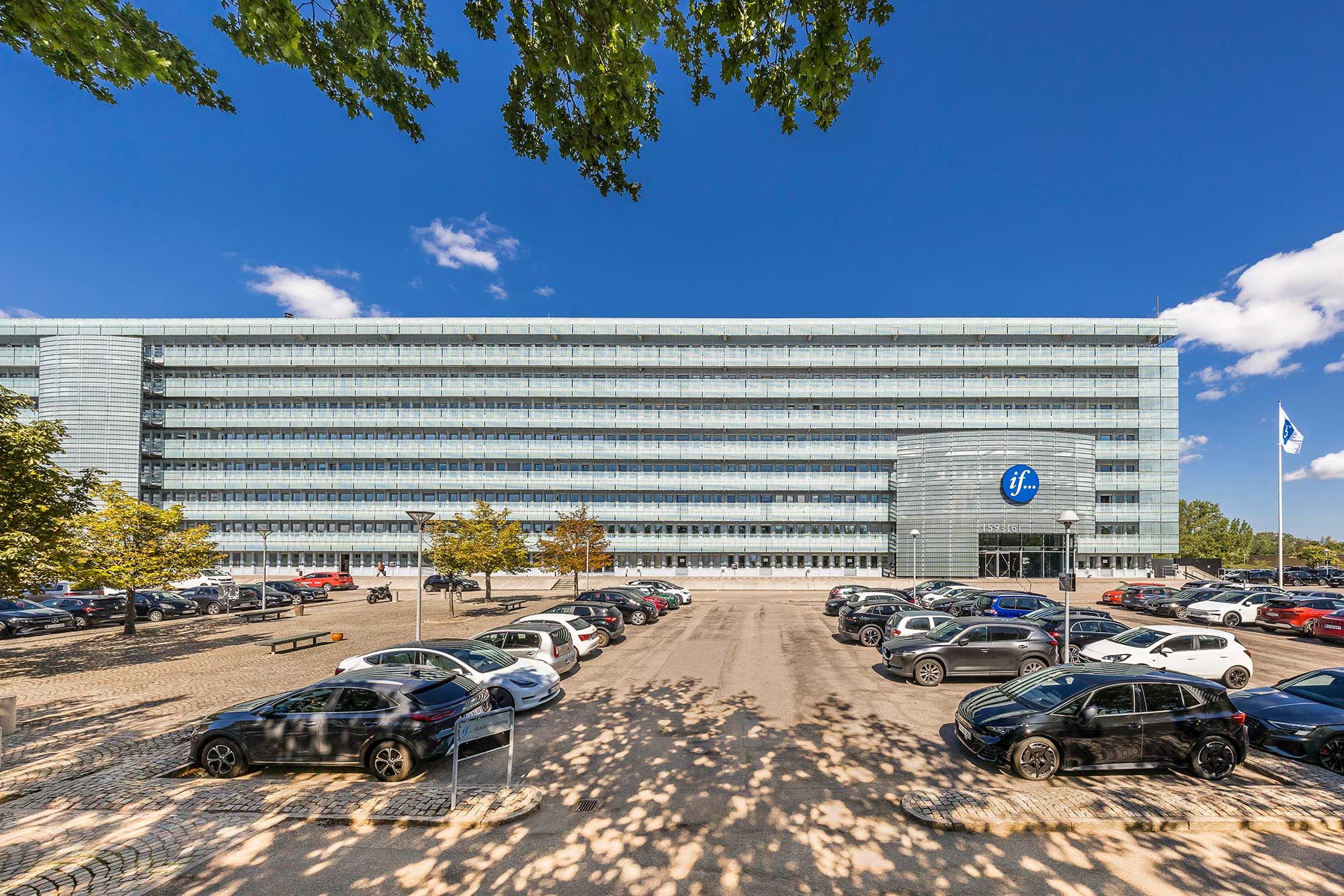 Unik kontorejendom med moderne design, glasfacade og logo på bygningen, omgiven af parkeringsplads og biler