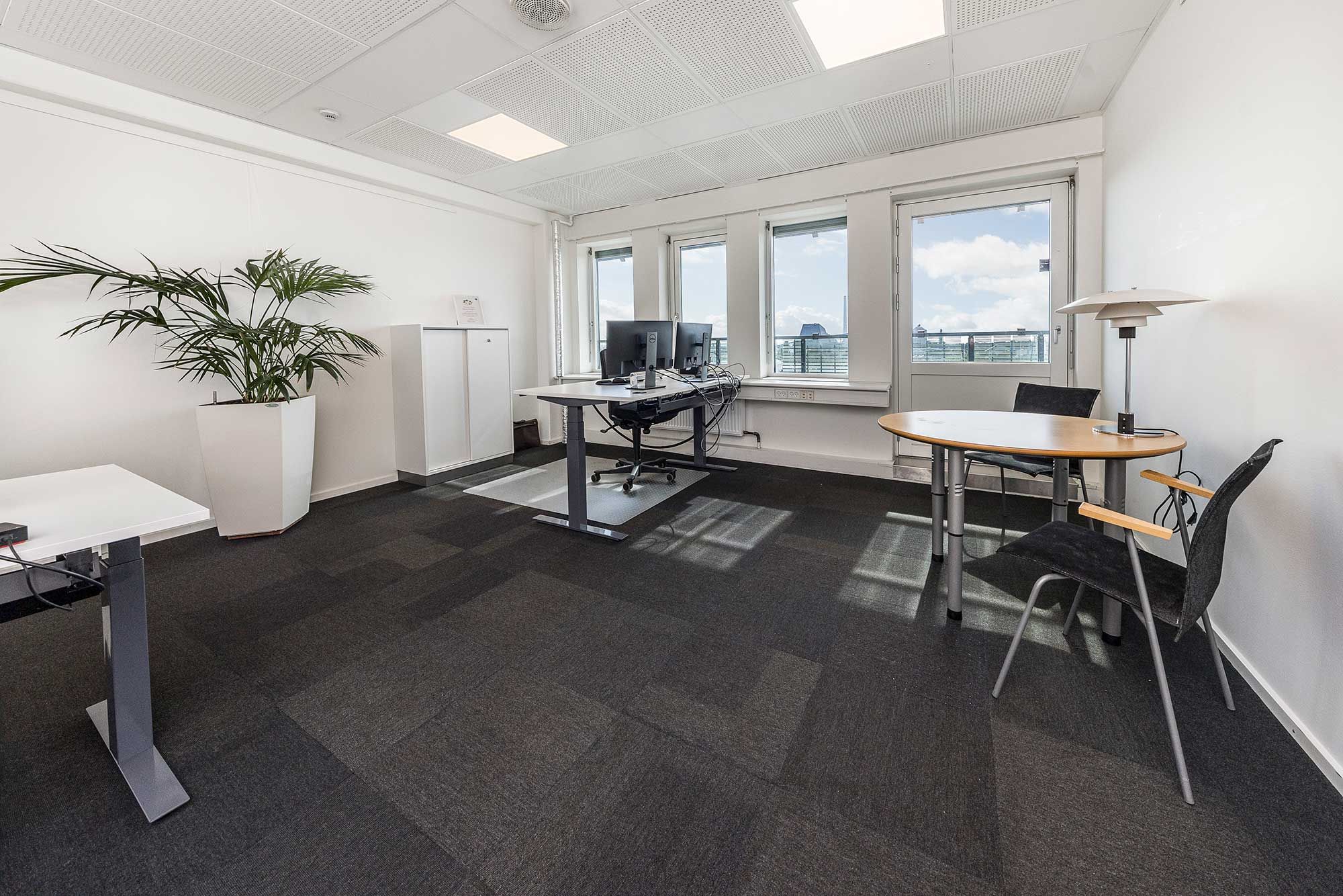 Kontor med fuld udsigt, højloft, lyse vinduer, moderne skrivebord, stol, rundt bord, plante, lampe og teknologiske faciliteter som computere og netværksstik.