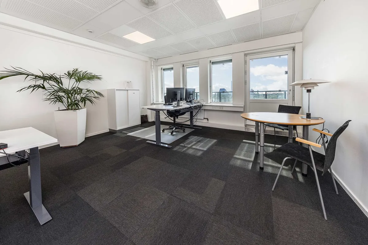 Kontor med fuld udsigt, højloft, lyse vinduer, moderne skrivebord, stol, rundt bord, plante, lampe og teknologiske faciliteter som computere og netværksstik.