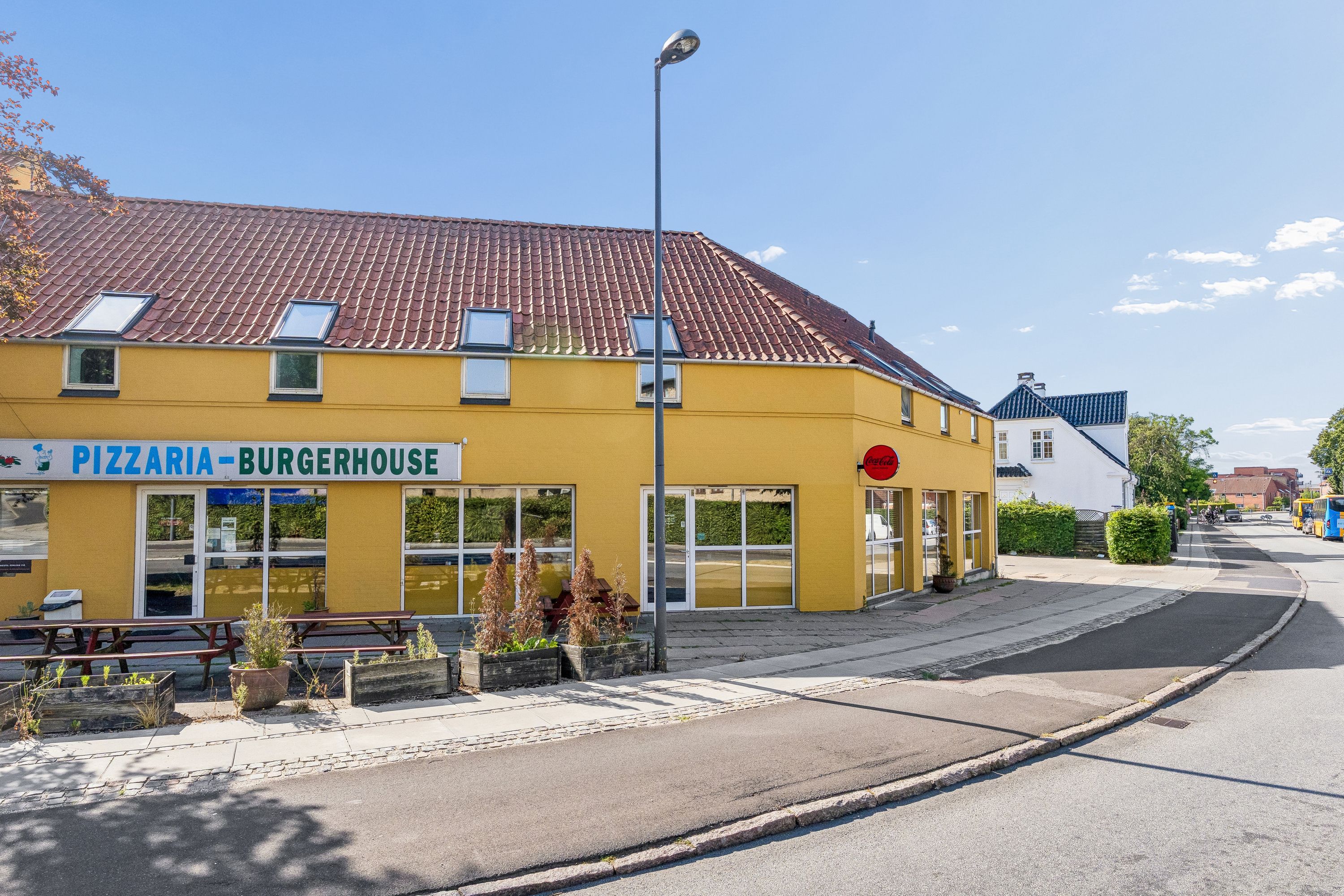 Pizzaria og Burger House med Coca Cola skilt uden for, lysegul facade, busser kører forbi og en slank streetlight i baggrunden
