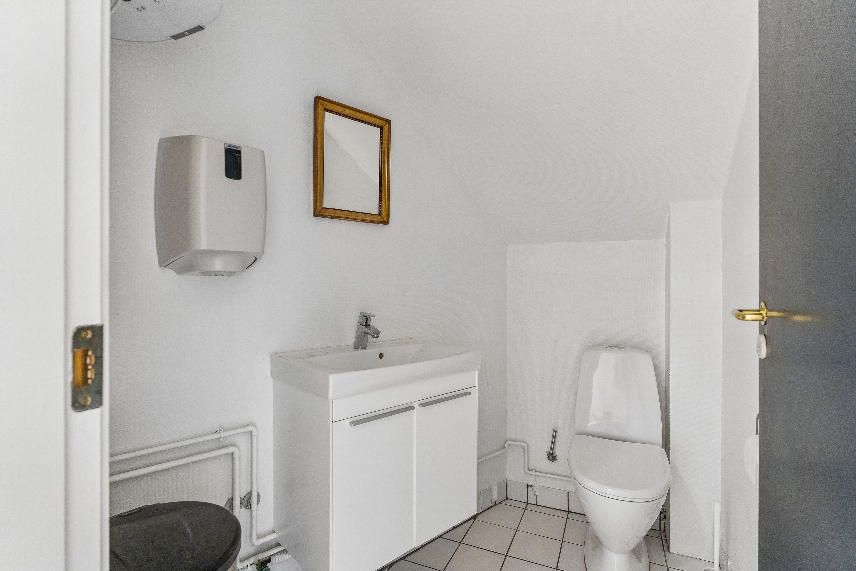 Rent toilet med hvidt tøj og toiletpapir ved siden af hængende spejl og håndklædetørrer. Toilet med lukket låg og hvidt porcelæn og hvidt gulv med hvide kakel.