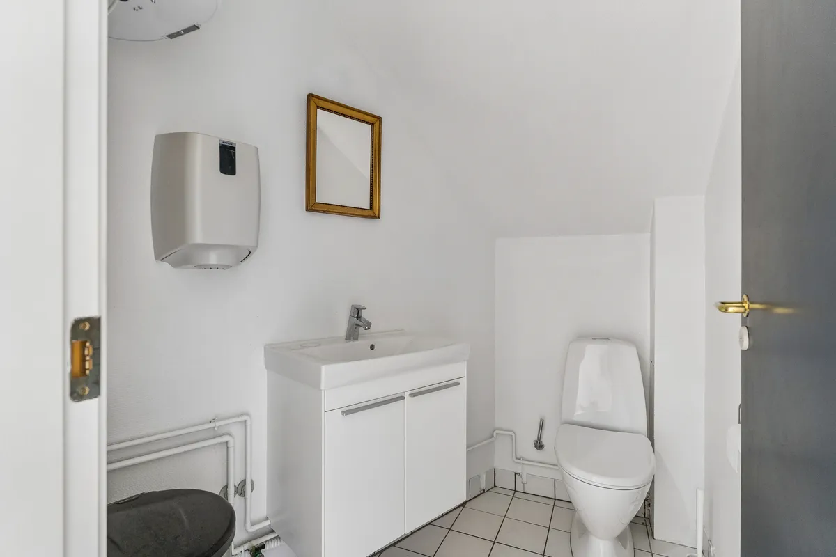 Rent toilet med hvidt tøj og toiletpapir ved siden af hængende spejl og håndklædetørrer. Toilet med lukket låg og hvidt porcelæn og hvidt gulv med hvide kakel.
