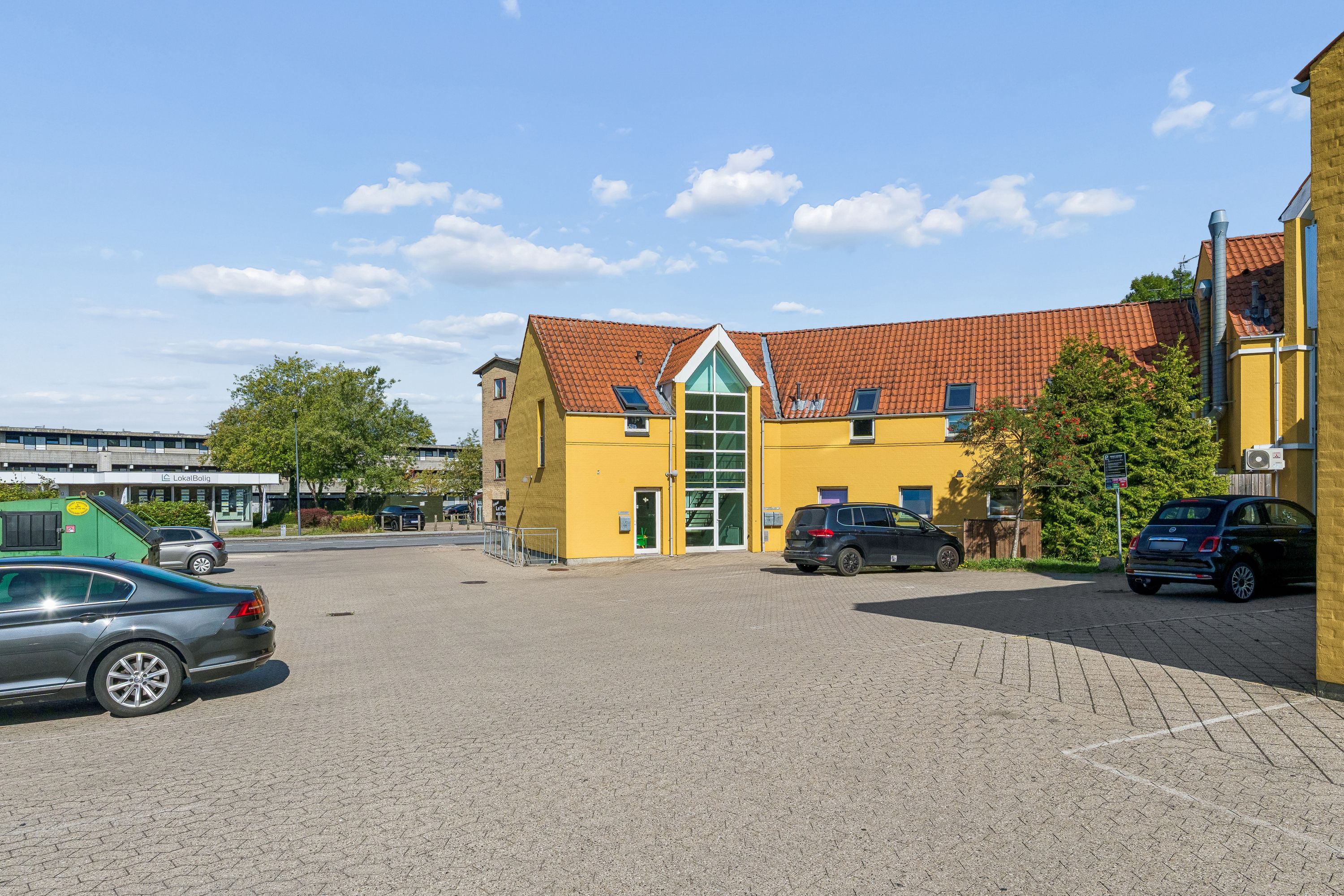 Et kontorlokal i Ballerup med gule vægge og rødt tag med to parkerede biler og et grønt affaldscontainer
