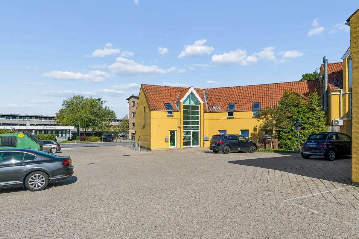 Et kontorlokal i Ballerup med gule vægge og rødt tag med to parkerede biler og et grønt affaldscontainer