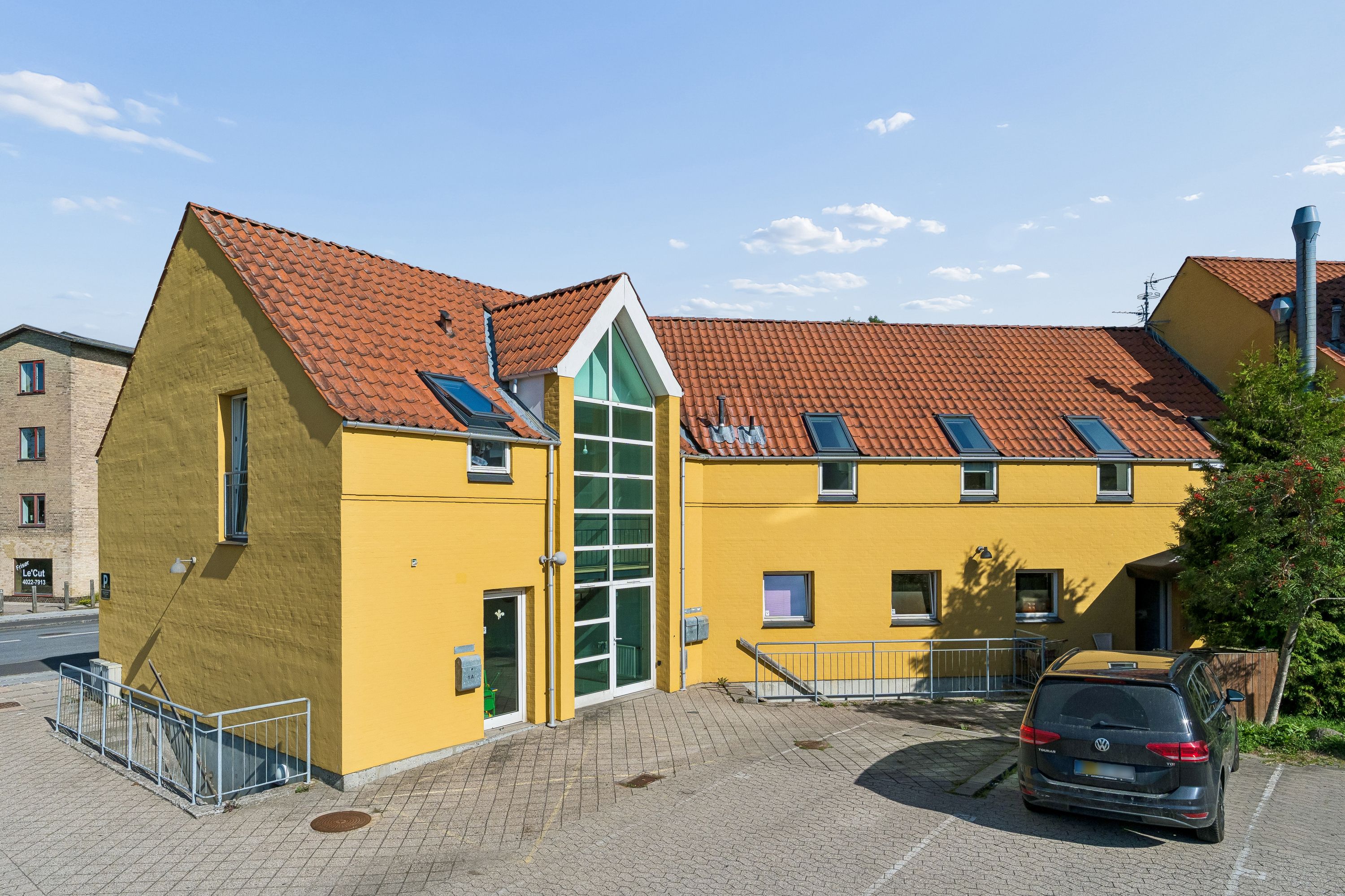 Kontorbyggeri med lysegul facade og rødt tag ligger i Ballerup. Der er solpaneler på taget, og parkeringspladserne er tæt ved. Et køretøj er parkeret foran bygningen