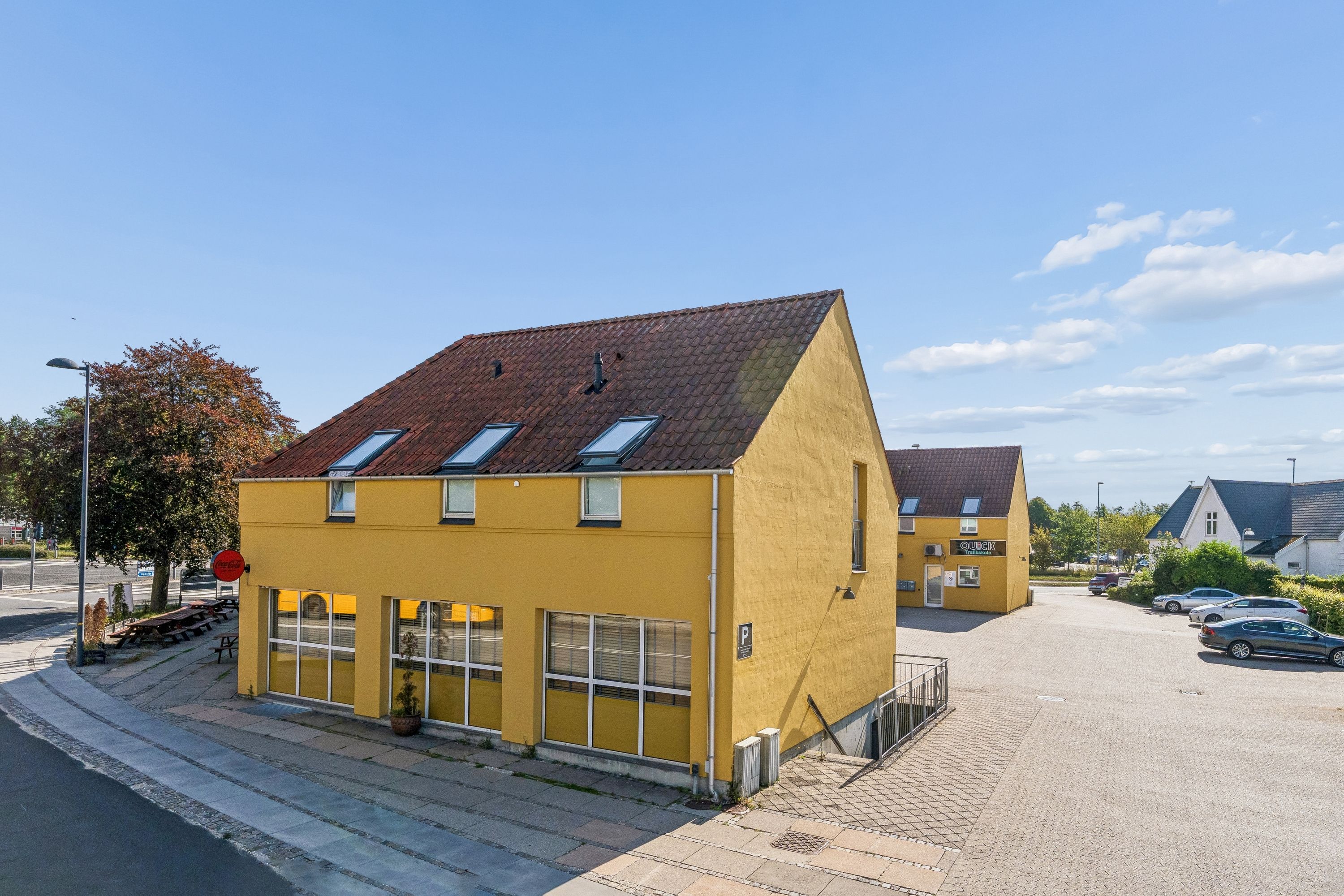 Lejlighedsbygning med gul facade og tegltag med Coca Cola-skilt. Lyst parkeringsområde med parkerede biler og udsigt mod en grønt bevoksket bakke.