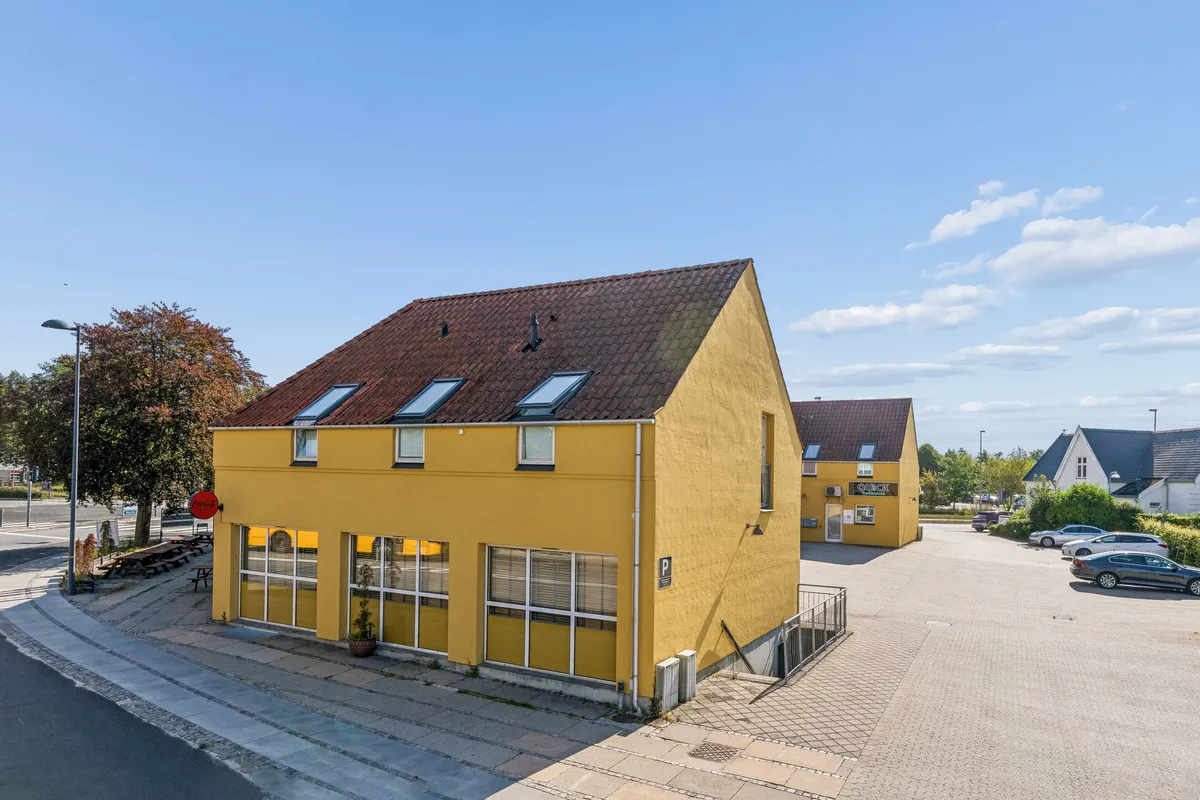 Lejlighedsbygning med gul facade og tegltag med Coca Cola-skilt. Lyst parkeringsområde med parkerede biler og udsigt mod en grønt bevoksket bakke.