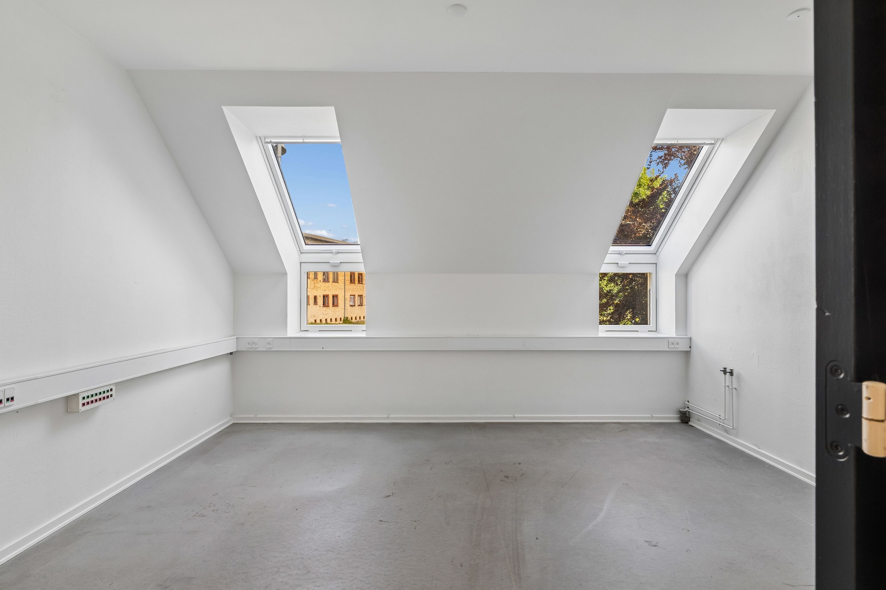 To loftsvinduer med udsigt til en bygning med røde mursten og lysegul facade. Væggene og gulvet er hvide og lyse.