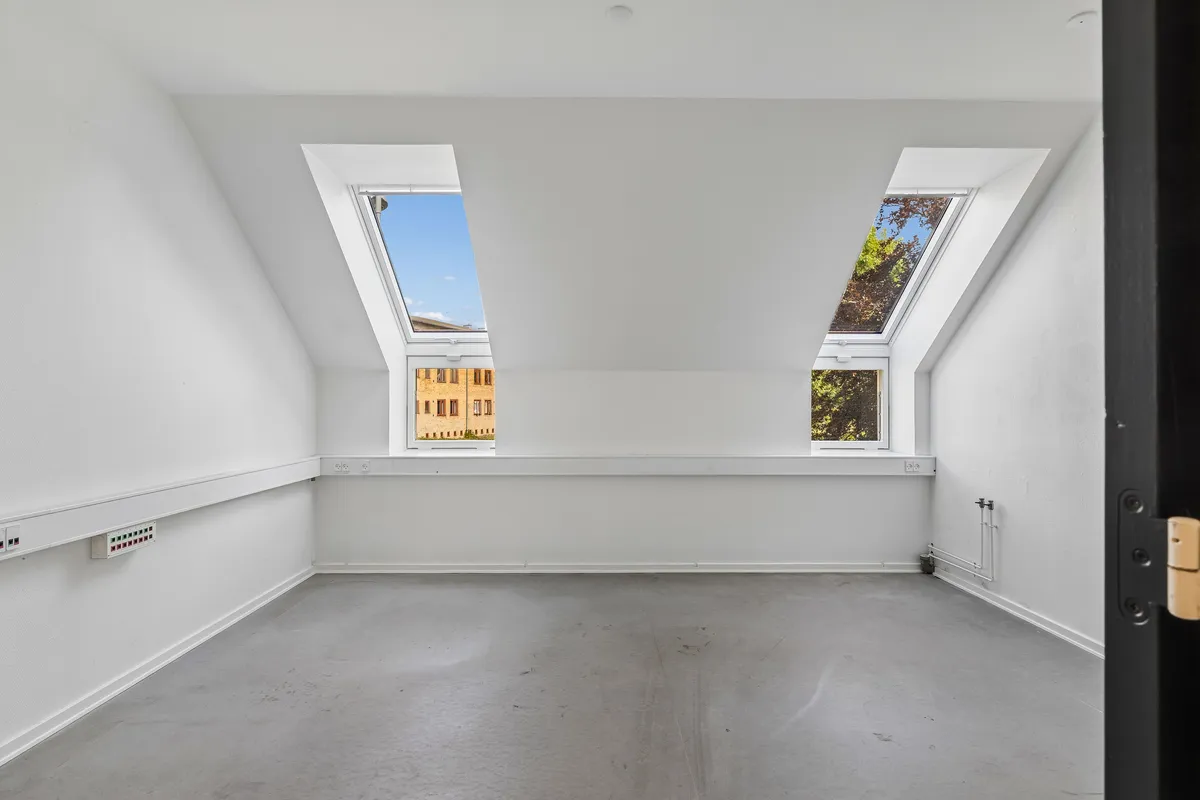 To loftsvinduer med udsigt til en bygning med røde mursten og lysegul facade. Væggene og gulvet er hvide og lyse.