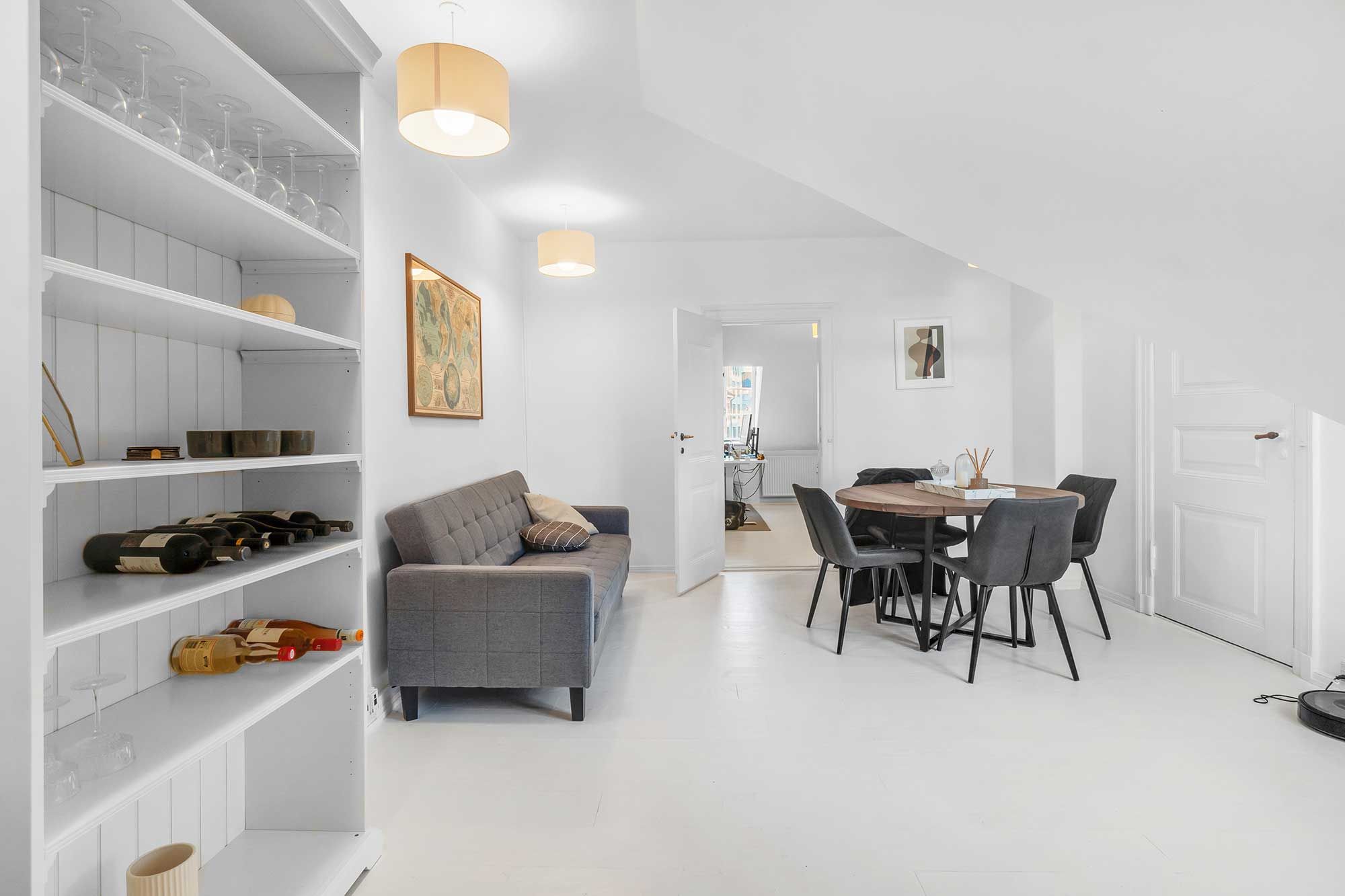 Hvidt interiør med højloft og sofa med puder, spisestue med bord og stole, lysekroner og hylder med vinflasker og glas