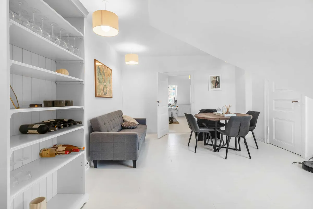 Hvidt interiør med højloft og sofa med puder, spisestue med bord og stole, lysekroner og hylder med vinflasker og glas