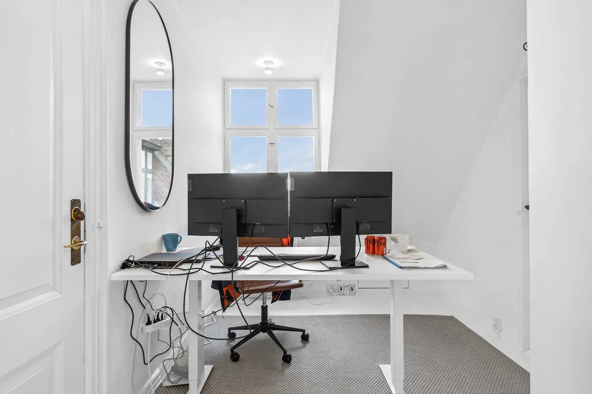 Kontorrum med skrivebord, dobbelt skærm, to kuglepenne og en kop kaffe på bordet og udsigt gennem vinduet mod Nytorv, Gammel Torv og Københavns Byret.
