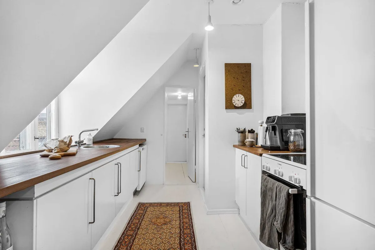 En lille køkken med hvide vægge og trægulv, integreret ovn og komfur, og et slankt design med moderne apparater og pæne indretningsgenstande