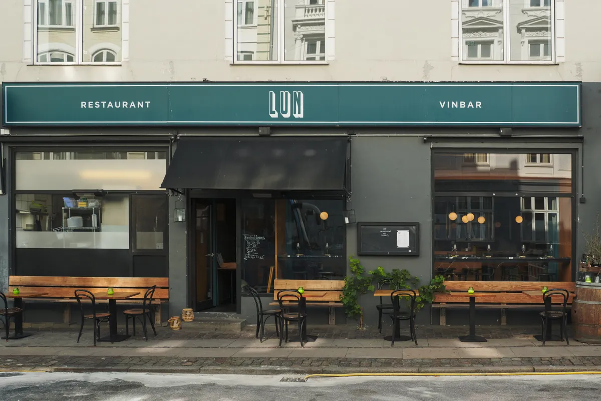 Udsigt til restaurant med lyse vinduer og indrettede bordsætninger. Grøn facade med restaurant og vinbar, lyse lamper og bordarrangementer. Bordsætninger med stole og bænke.
