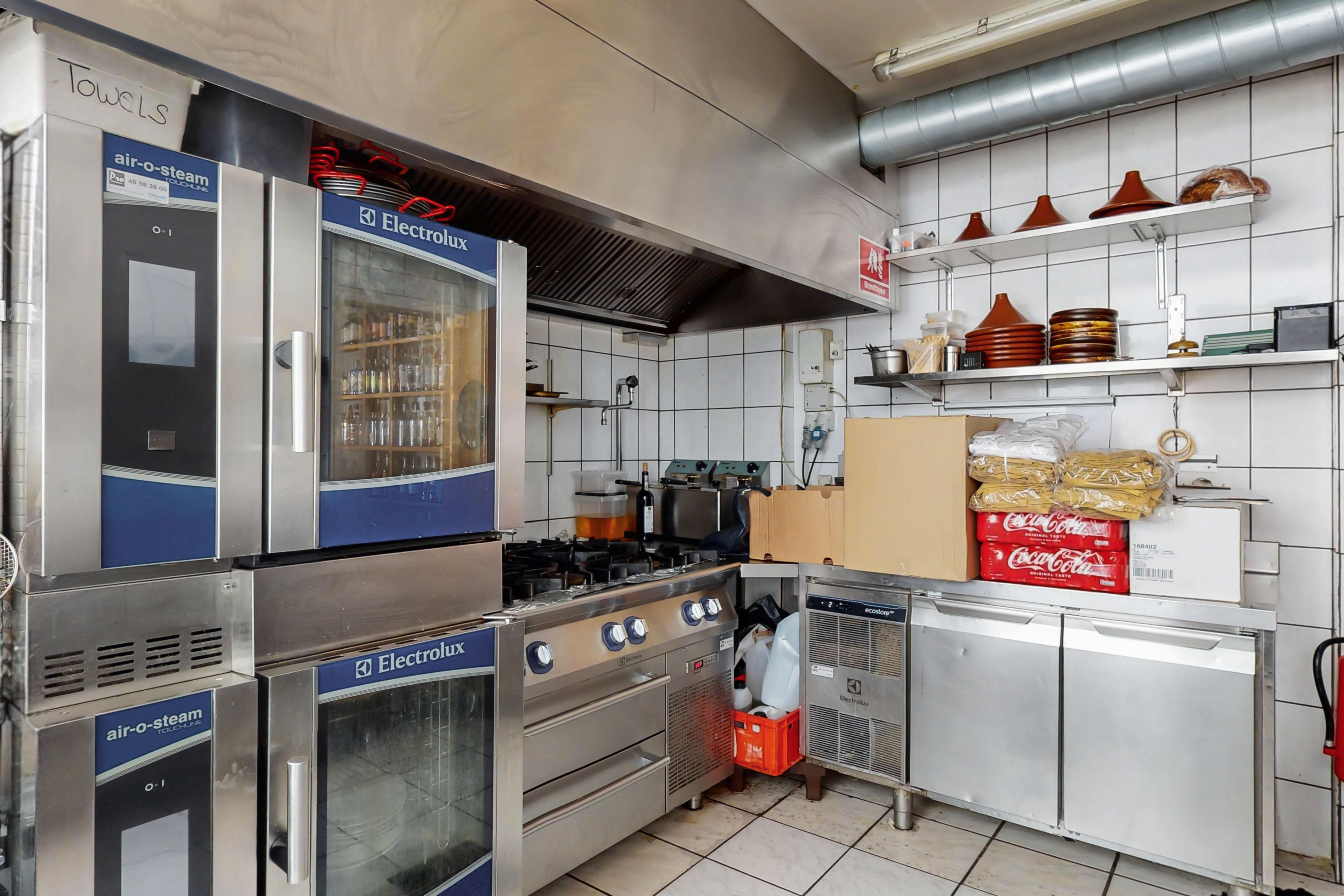 Moderne kommercielt køkken med store apparater, herunder Electrolux ovne og kogeapparater, og lagerhylder med diverse emballage og tøj.