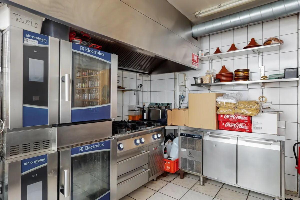 Moderne kommercielt køkken med store apparater, herunder Electrolux ovne og kogeapparater, og lagerhylder med diverse emballage og tøj.