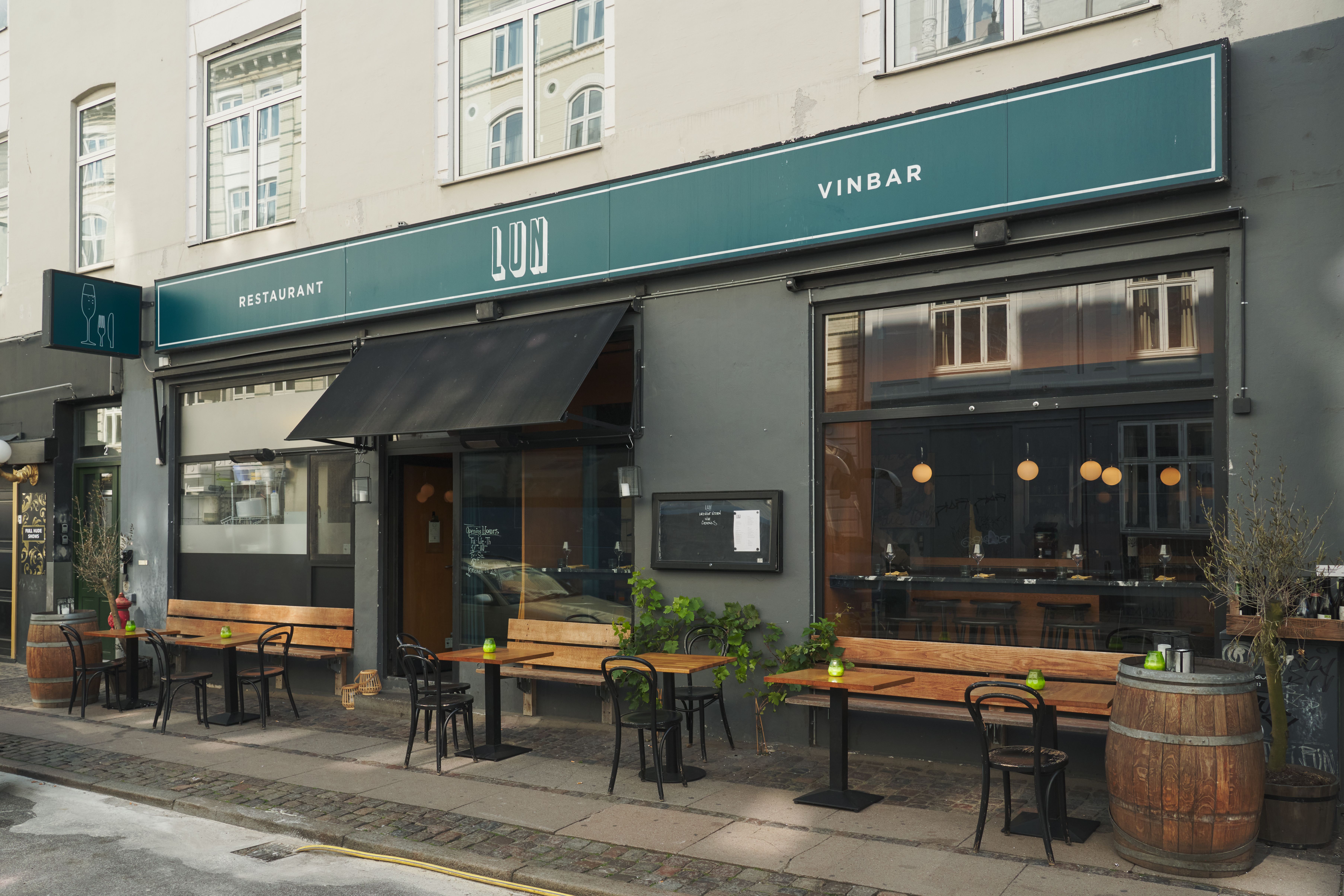 En vinbar med navnet Vinbar Lun ligger på Helgolandsgade 2, 1653 København V. Der er stole og borde udenfor og et træfælde. Der er plante i lysekroner og en træbænk med et vinglas på bordet.