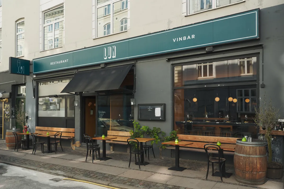 En vinbar med navnet Vinbar Lun ligger på Helgolandsgade 2, 1653 København V. Der er stole og borde udenfor og et træfælde. Der er plante i lysekroner og en træbænk med et vinglas på bordet.