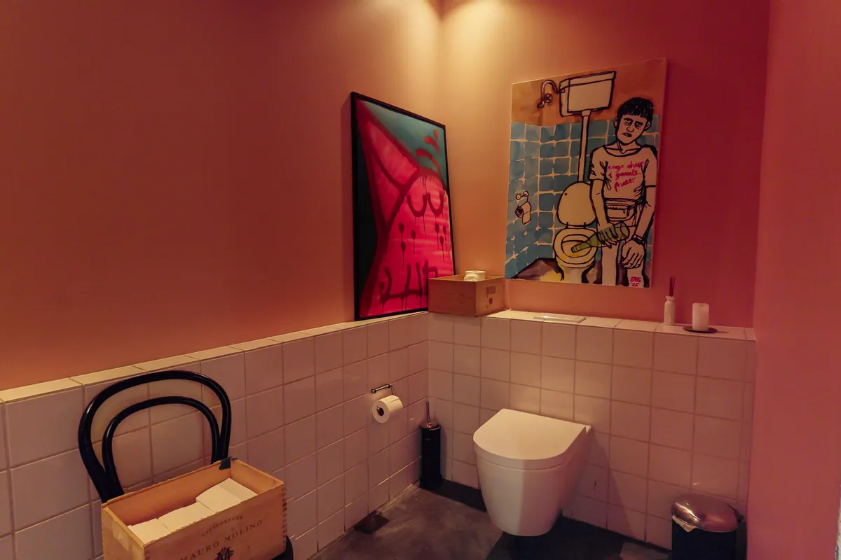 Toilet med toiletbørste, toiletpapir, håndklæder, kunstværker og tøjstang i lys pink badeværelse med trævinkasse og tøjstang