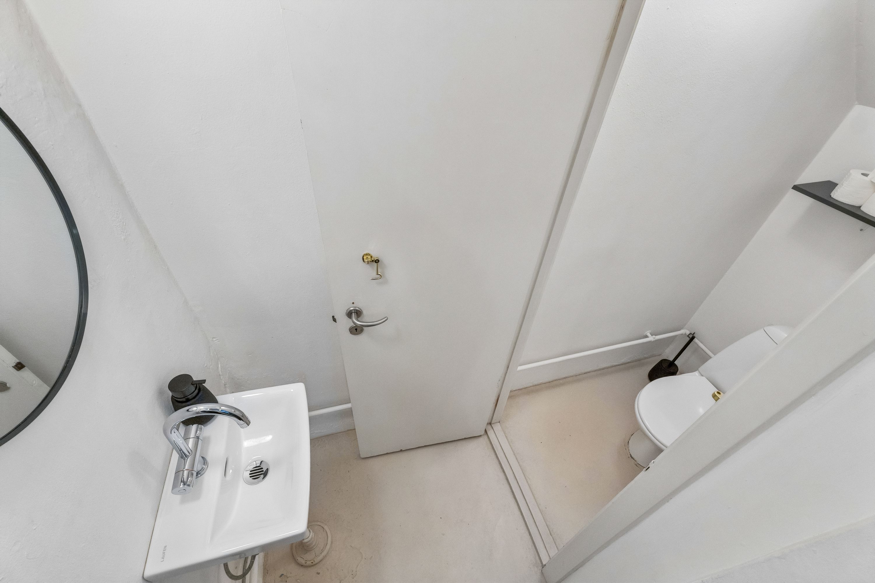 En lille toiletrum med hvidt toilet, håndvask med kromfarvet vandhane, spejl og toiletpapirhylde samt et håndstik på væggen