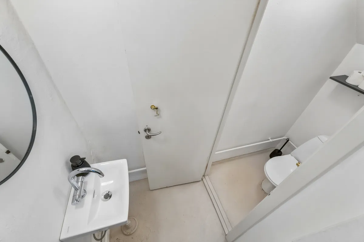 En lille toiletrum med hvidt toilet, håndvask med kromfarvet vandhane, spejl og toiletpapirhylde samt et håndstik på væggen