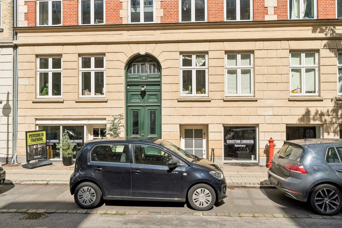 To biler parkeret på gaden, en mørk og en grå. Et rødbrandsslukker og en plante i potter. Lejlighedsdøre med nummeret 8 og Sebastian Lundberg Diplomtræner.