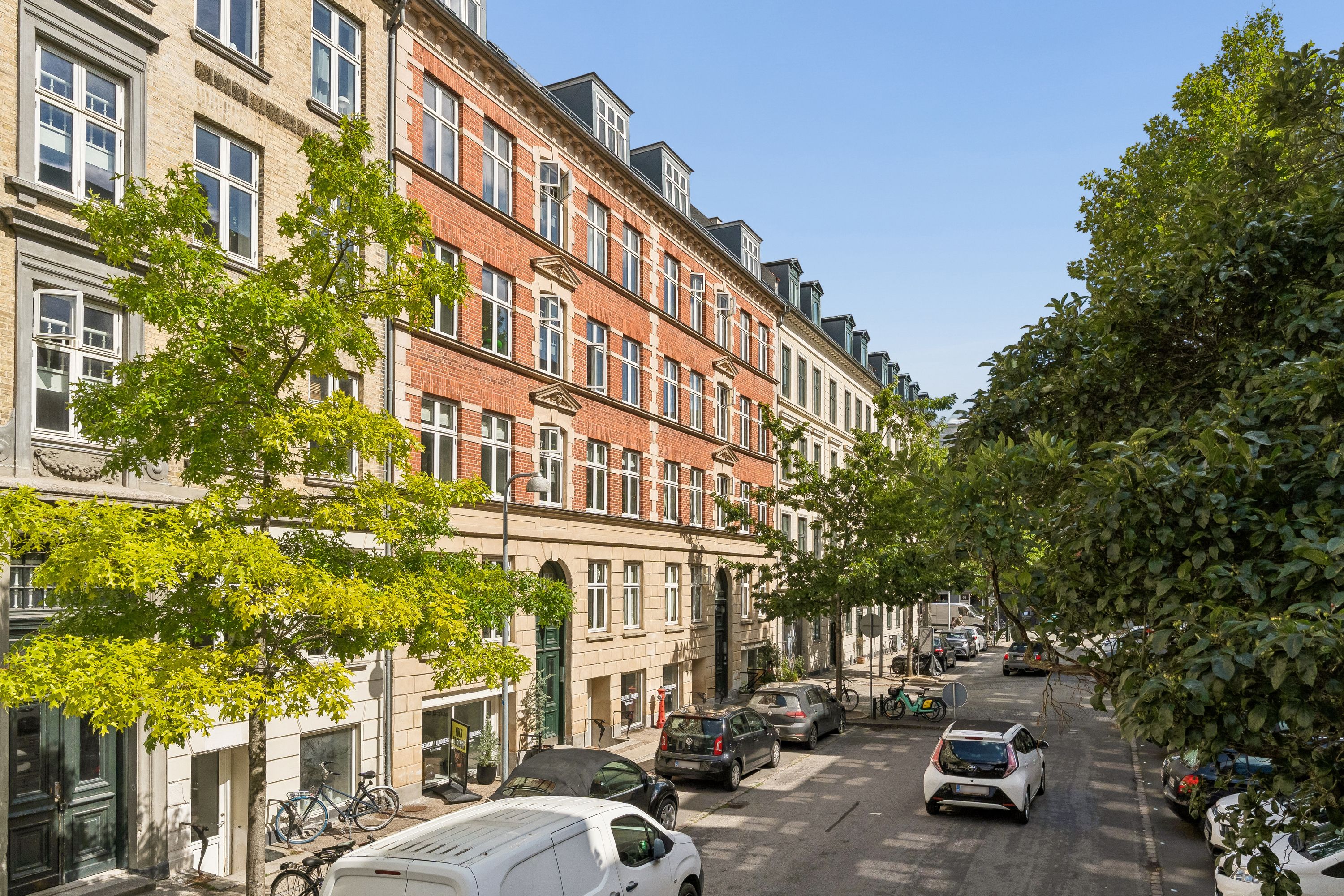 Kontor til leje i Frederiksberg. En række bygninger med røde mursten og hvide vindueskarme. Parkerede biler og cykler langs gaden. Solrig dag med højt træ og buske på begge sider