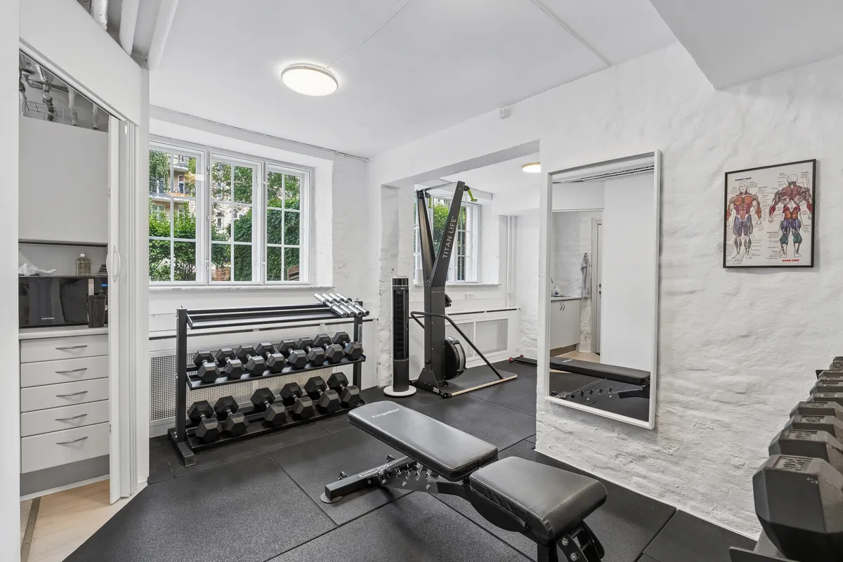 Moderne hjemmetræningsværelse med højloft, fitnessudstyr, sammenklappelig bench, stang med veje og sæt dumbbells, trægulv og lyse vinduer med udsigt til gården
