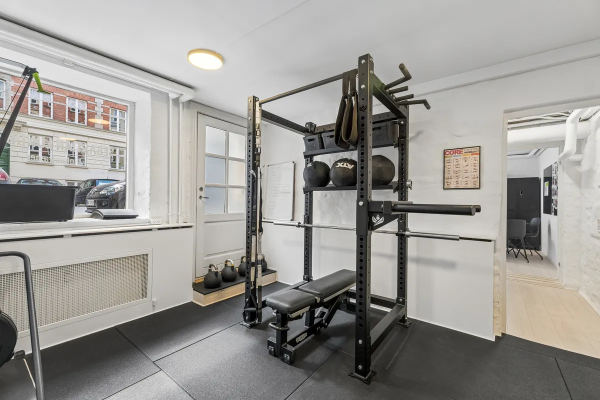Lejlighed med luksuriøs fitnessrumindretning. Sort styrketræningsmaskine med kettlebells og vægtkløver. Rådgivende træningsposter og skrifttavle. Lys og moderne interiør med trægulve og lyse hvide vægge